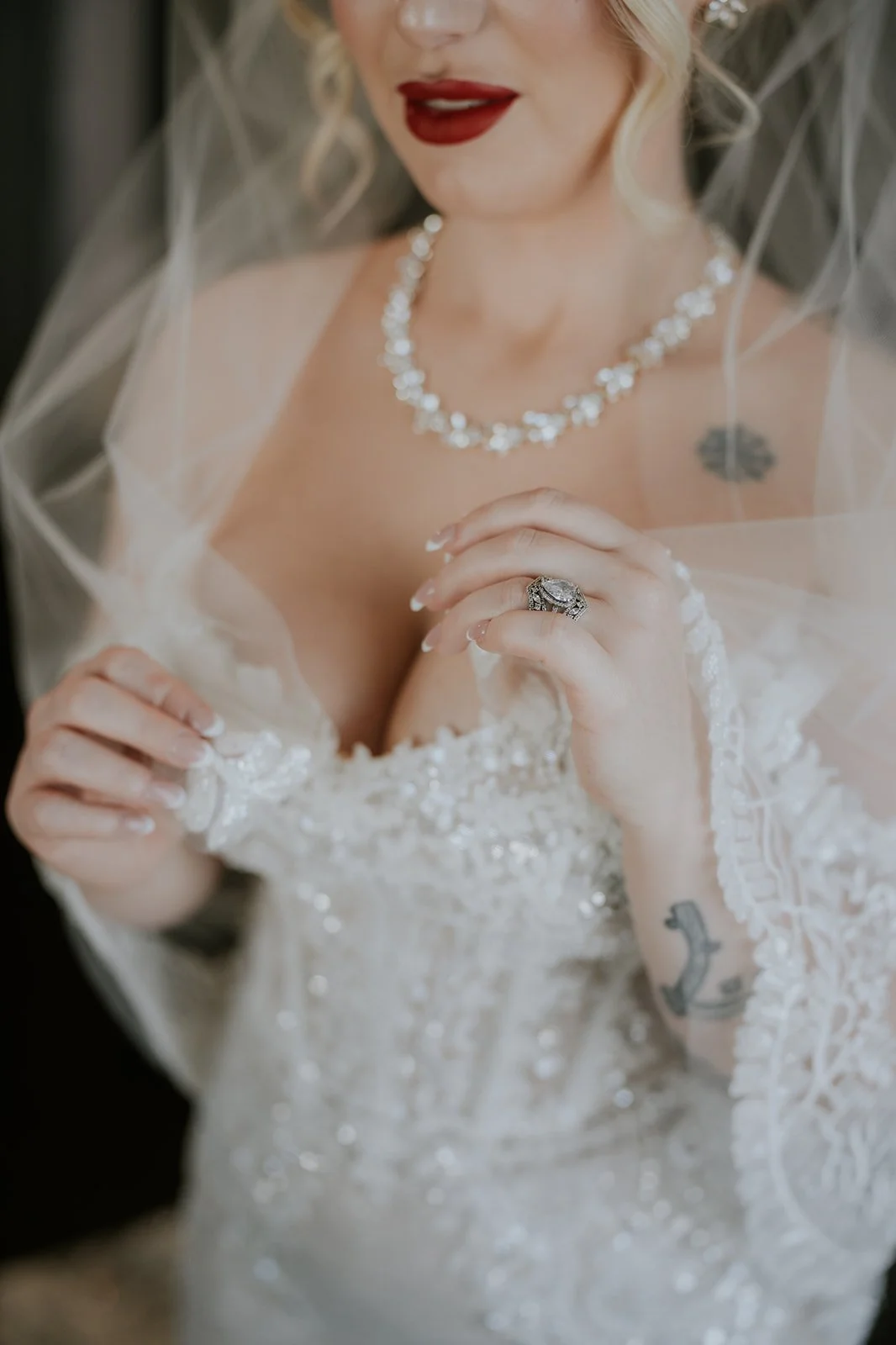 Close-up of a bride wearing a wedding dress with lace and bead details, a necklace, red lipstick, and a large diamond ring on her finger, partially covered by a sheer veil.