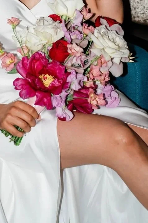 A person holding a bouquet of assorted colorful flowers, including pink, white, and red blooms, while sitting with legs crossed on a white surface.