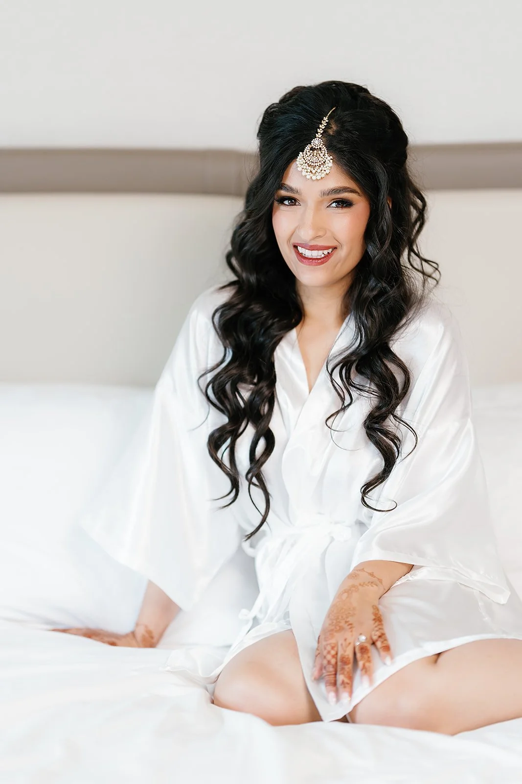 Trusha seated on bed in bridal robe with maang tikka and henna during getting ready at JW Marriott Las Vegas wedding, hair by Courtney and makeup by Natalia for Amelia C & Co, photography by Chernogorov Photography