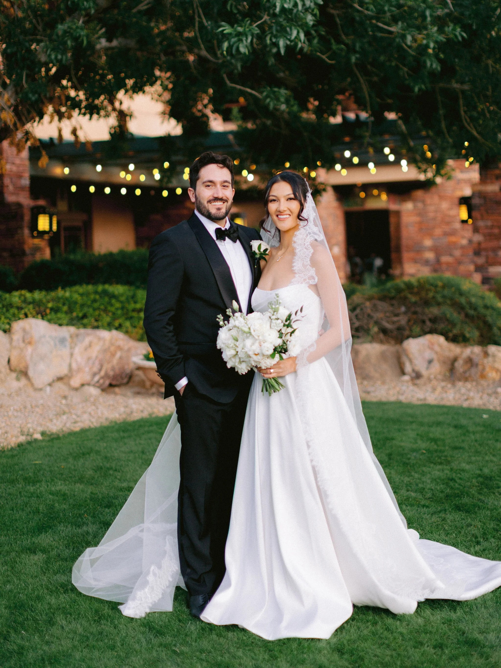 Bride and groom portrait at golden hour at Las Vegas Paiute Golf Resort wedding with string lights backdrop and full cathedral veil, hair by Kari K and makeup by Natalia W for Amelia C & Co, photography by Susie and Will