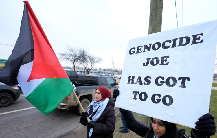Two women participate in a protest; one holds a Palestinian flag, the other holds a sign that reads 'Genocide Joe Has Got To Go'.