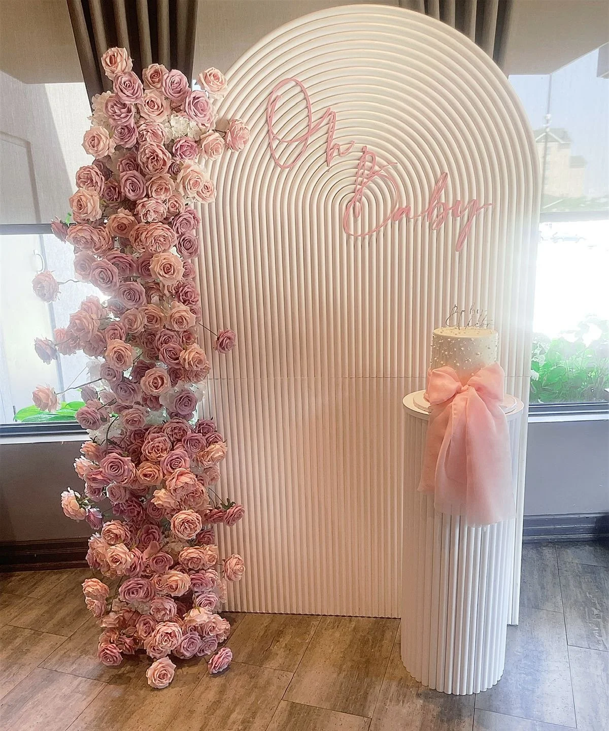 A decorative setup for a celebration with a backdrop featuring a vertical, ribbed white panel, a cascading arrangement of pink and peach roses on the left, a framed pink neon sign that says 'Amor' in the center, and a white pedestal on the right with