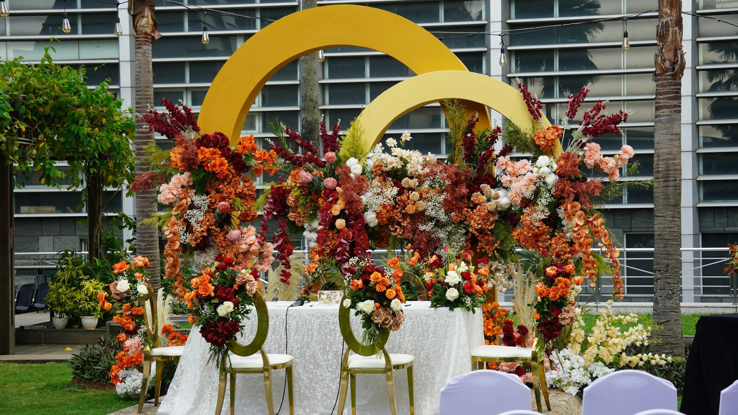 A floral wedding arch decorated with various colorful flowers, set up outdoors with chairs and a white tablecloth beneath it, in front of a modern building with glass windows and palm trees.