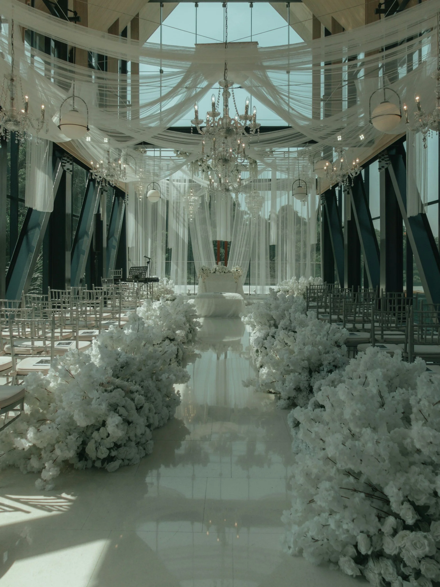 Elegant indoor wedding ceremony setup with white floral aisle, draped fabric and chandeliers.