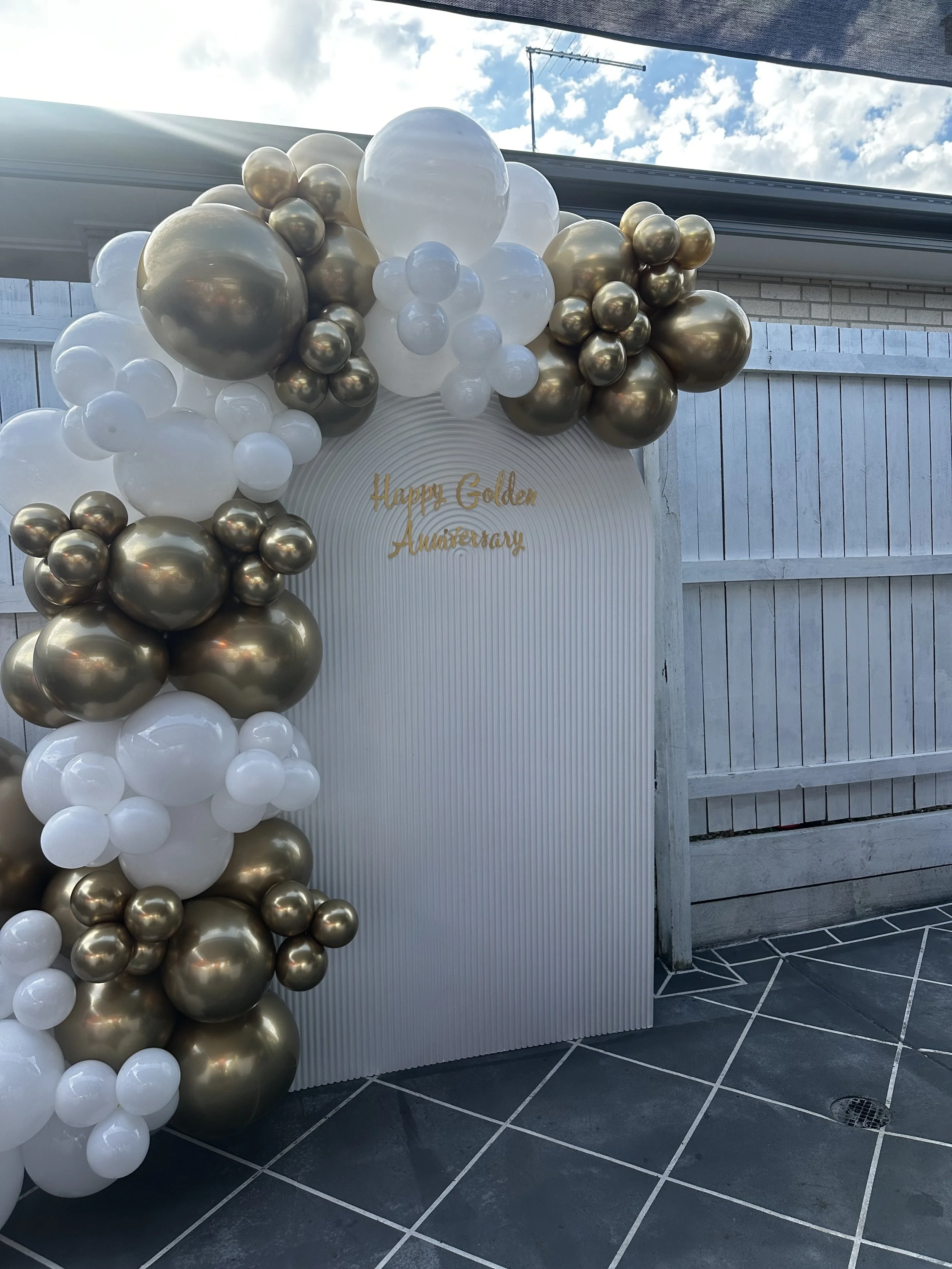 Balloon arch with white, gold, and metallic silver balloons, and a backdrop with gold script reading 'Happy Golden Anniversary'.