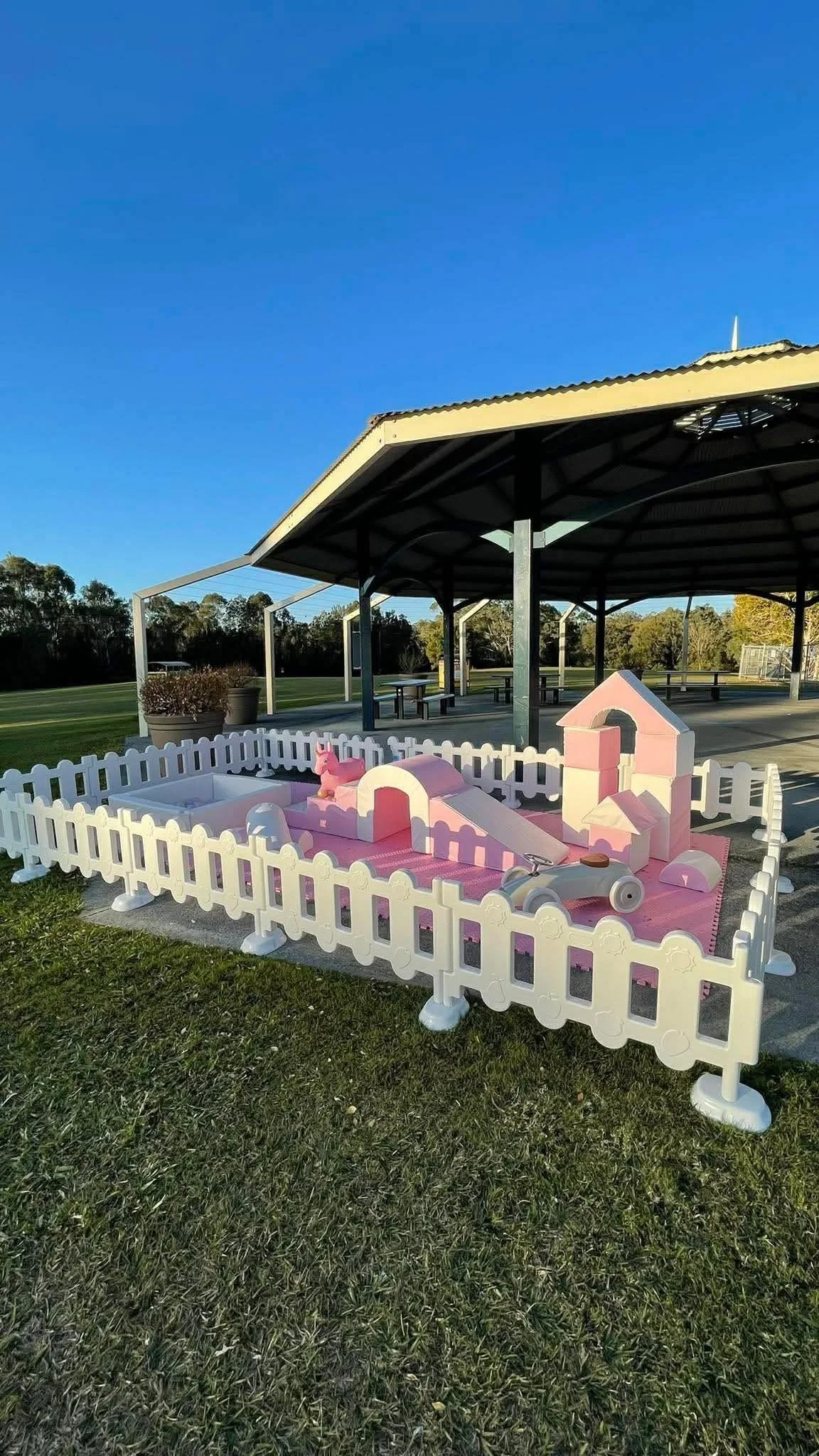 A pink and white plastic playpen with a playground set and a small slide, situated on a grassy field near a covered pavilion with benches and tables, under a clear blue sky.