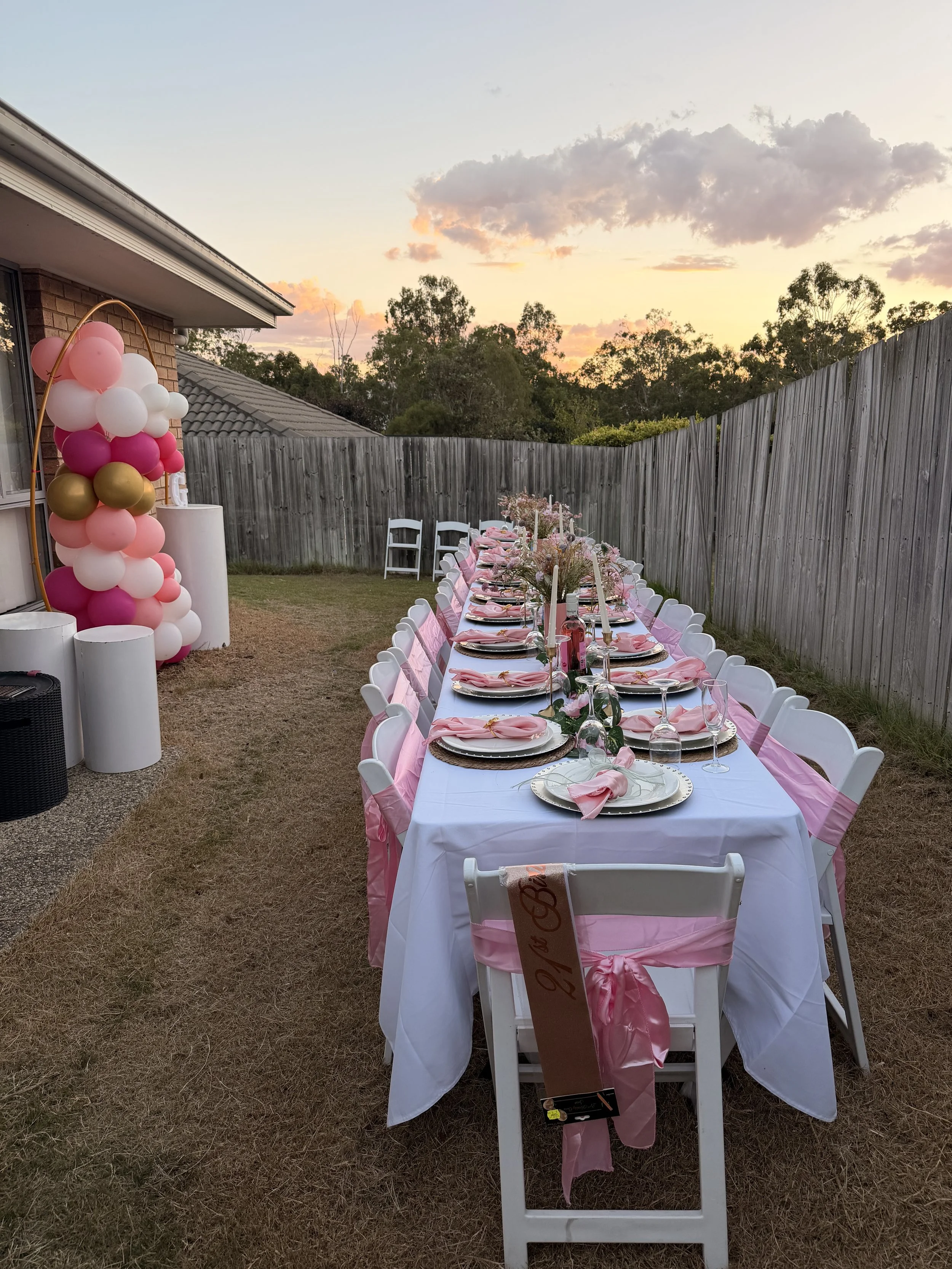 Outdoor party set up with a long table dressed in white with pink accents, decorated with flowers, candles, and glassware. Chairs are adorned with pink sashes, and a balloon arch in pink, white, and gold is nearby. The setting is in a backyard with a