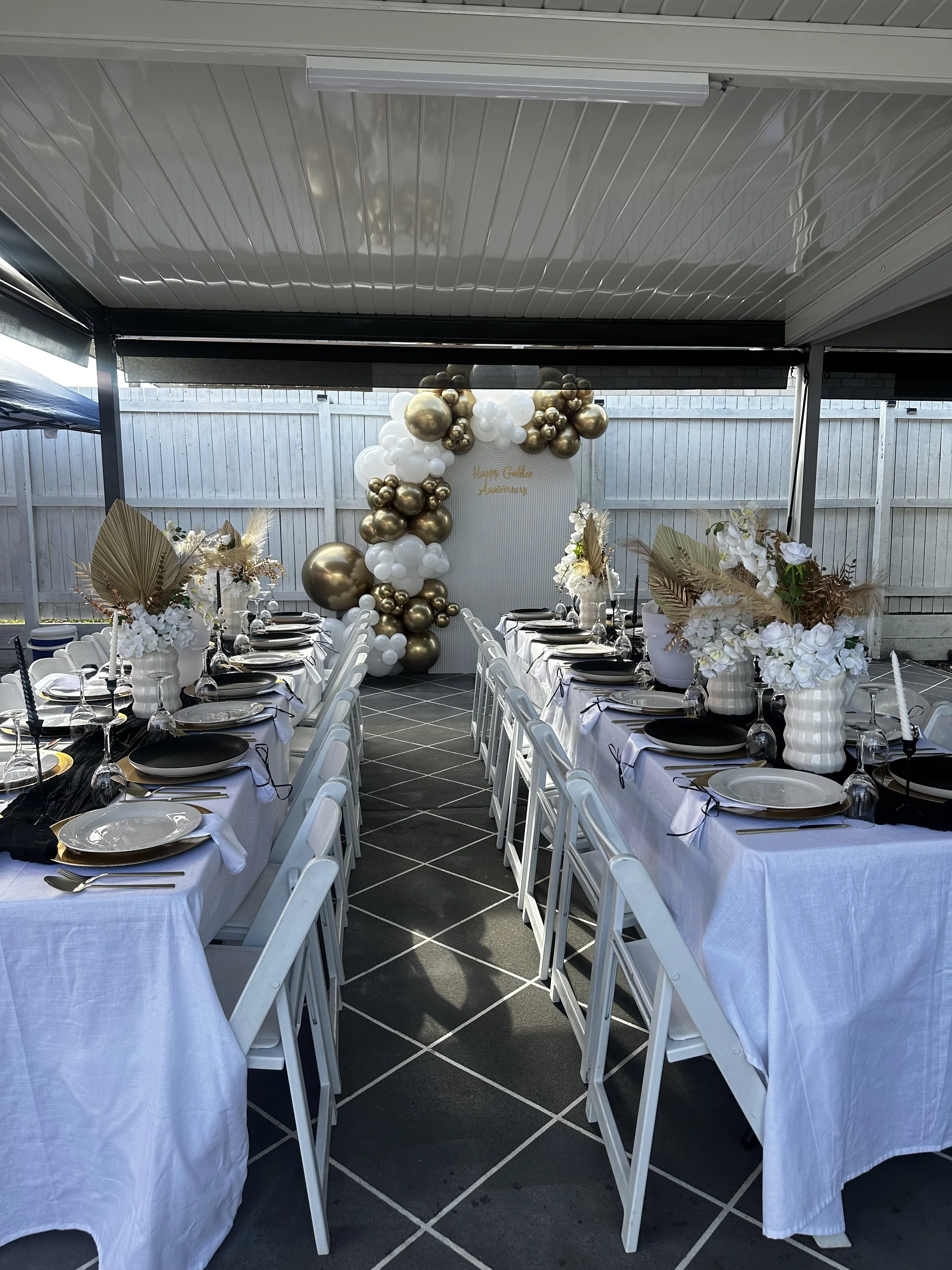Wedding reception setup with long tables covered in white tablecloths, decorated with black and gold accents, and surrounded by white chairs. There are floral arrangements in white vases and a balloon arch with white, gold, and metallic balloons behind a white backdrop with gold writing. The setting is in a covered outdoor area with a white wooden fence in the background.