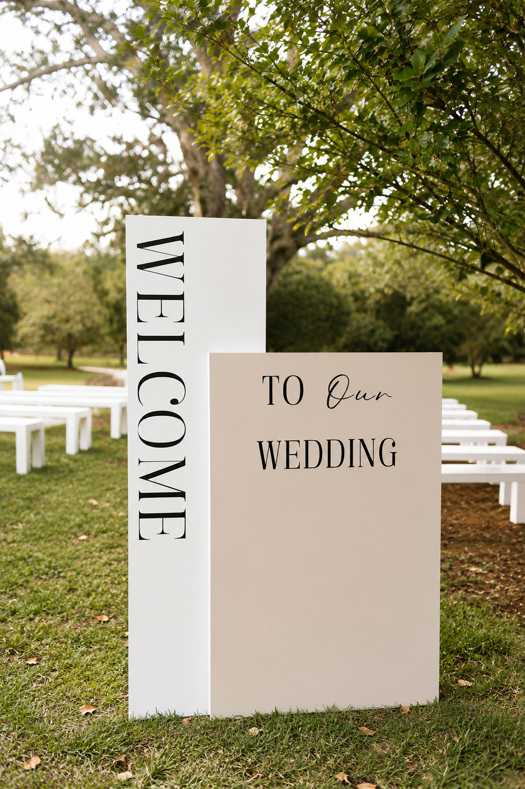 Outdoor wedding welcome sign with white and beige panels, readin "Welcome to Our Wedding," set on grassy area with white benches and trees in the background.