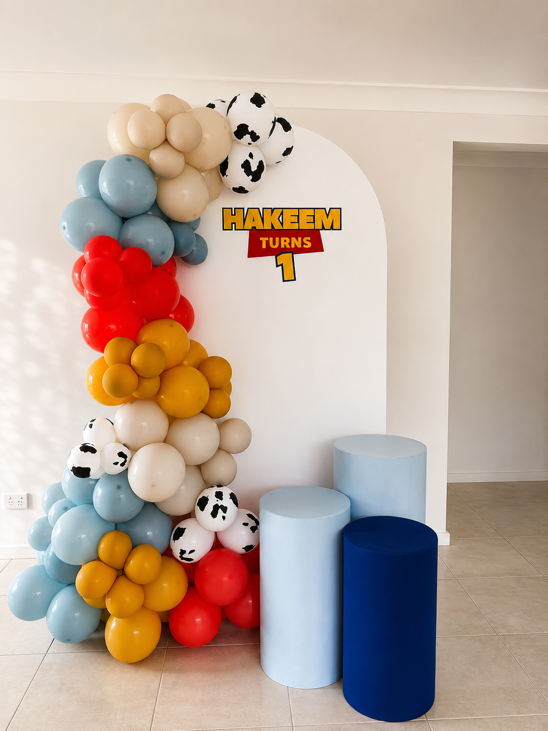 Colorful balloon arch with black and white cow-print balloons, and a sign that says "Hakeem Turns 1" in front of a white wall.