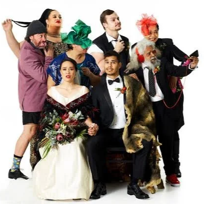 Group of seven diverse people in costumes, including a woman with a flower bouquet, a man in a tuxedo with a beard, a woman with bright green hair, a man in a suit, a woman with bright red hair and a phone, an elderly man with a Santa beard, and a dog wearing a fur coat.