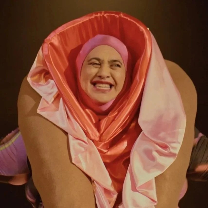 Person with a humorous facial expression wearing a pink head covering and wrapped in a pink and peach fabric, positioned within a heart-shaped frame.