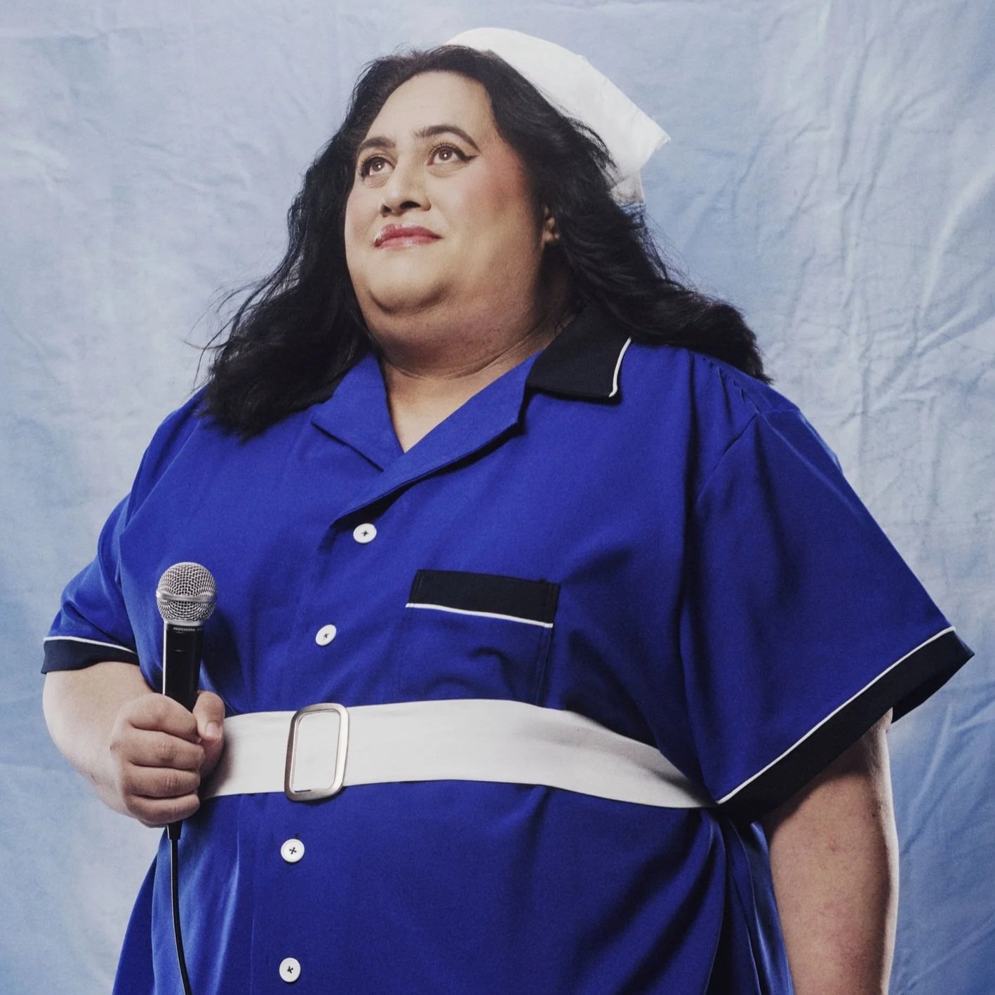 A plus-sized woman with long black hair, makeup, and eyeliner, wearing a blue dress with white piping and black accents, holding a microphone.