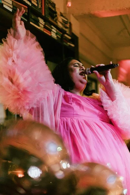 Woman singing passionately into a microphone, wearing a flowing pink dress with fluffy sleeves, in a cozy indoor setting with bookshelves in the background.
