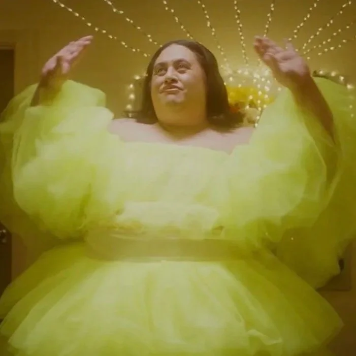 A plus-sized woman in a bright yellow dress with puffed sleeves, raising her arms with a smile, against a gold background with radiating light patterns.