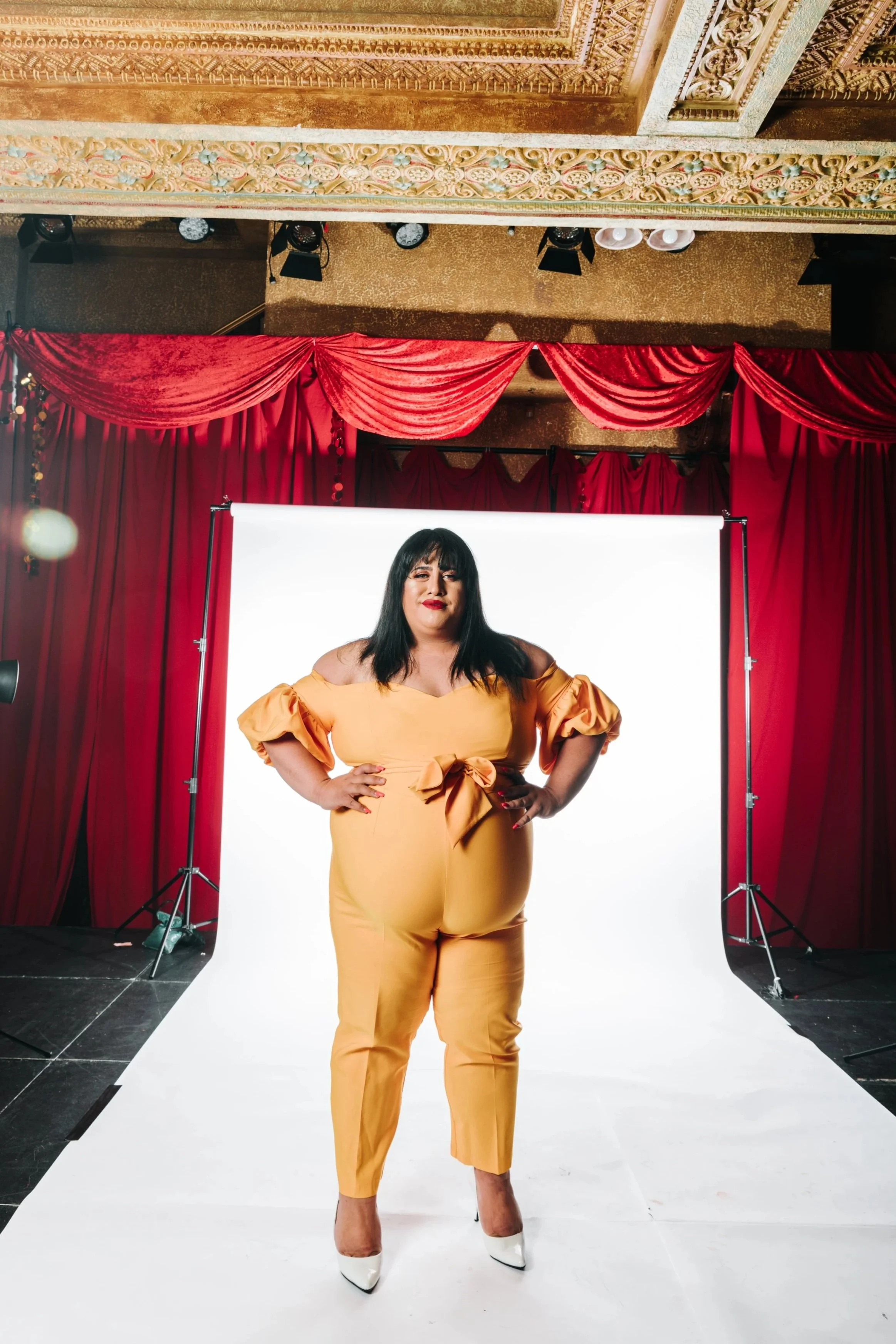 Woman wearing a yellow jumpsuit and white high heels standing in front of a white backdrop with red curtains in a photo studio.