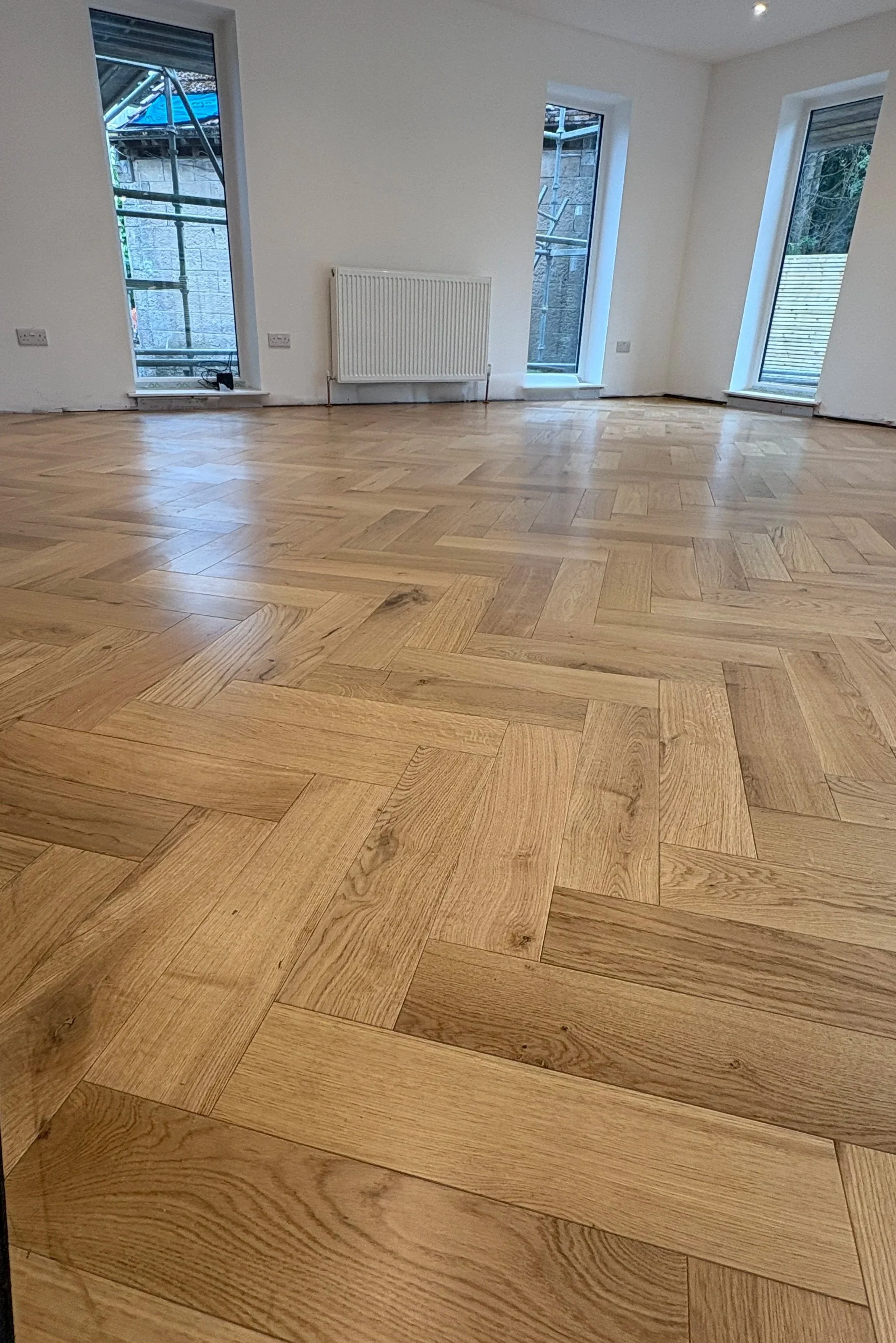 Empty room with three large windows, a white radiator, and a wooden herringbone-patterned floor.