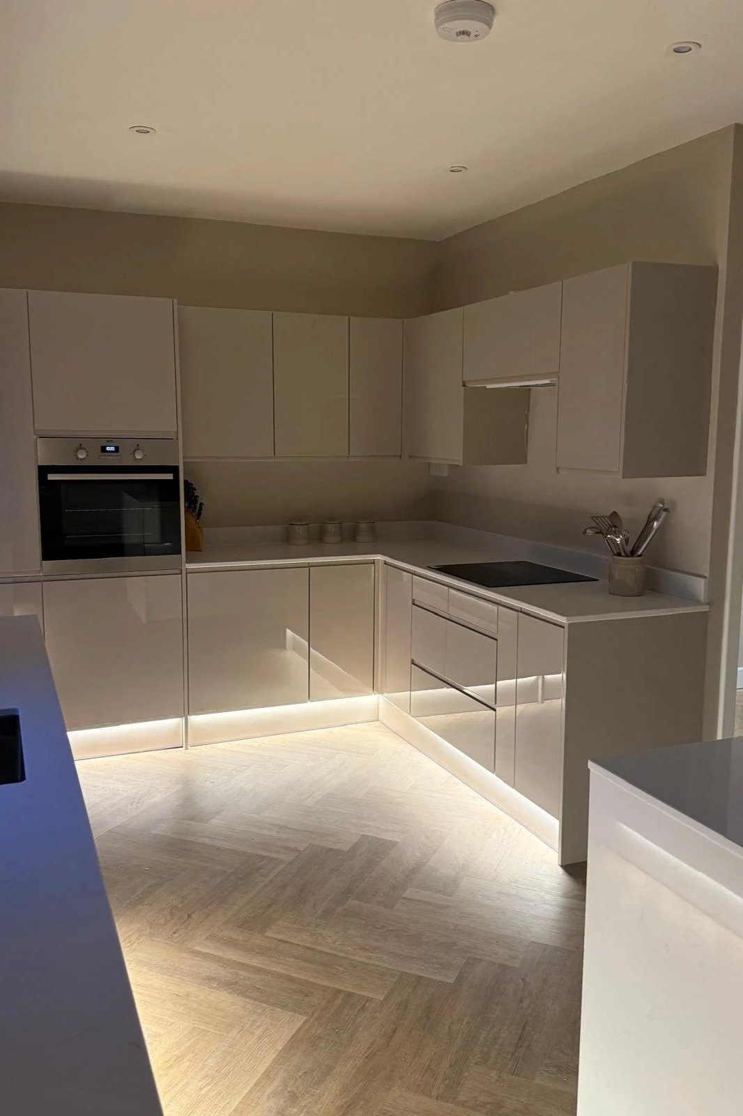 Modern kitchen with beige cabinets, white countertop, built-in oven, cooktop, utensils holder, and ambient lighting.