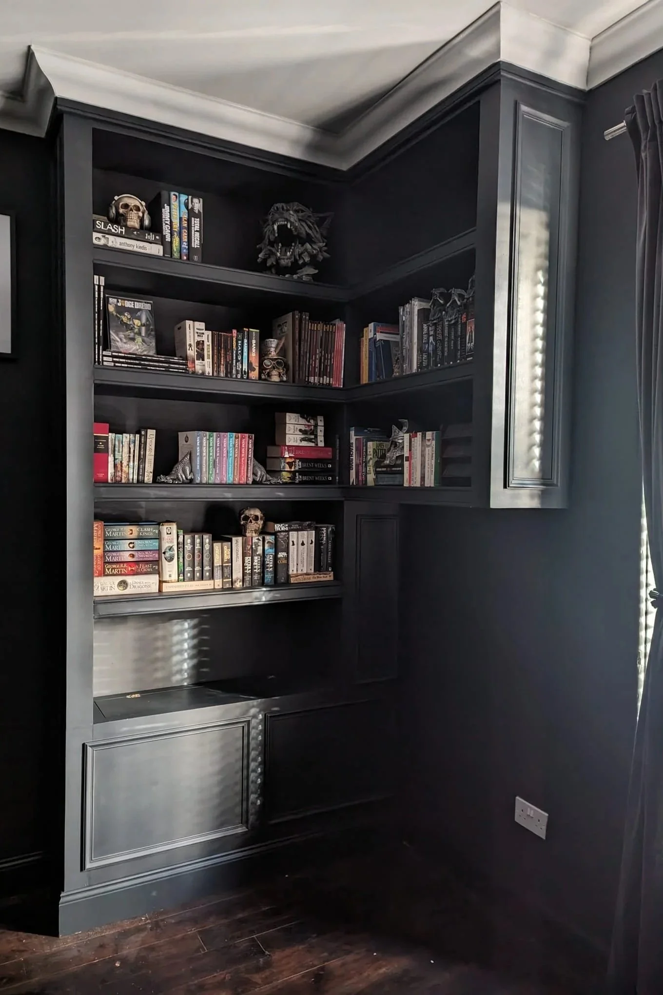 A black bookshelf against a dark wall, filled with books, decorative skulls, and a black lion head sculpture, with a dark floor and gray wall paneling.