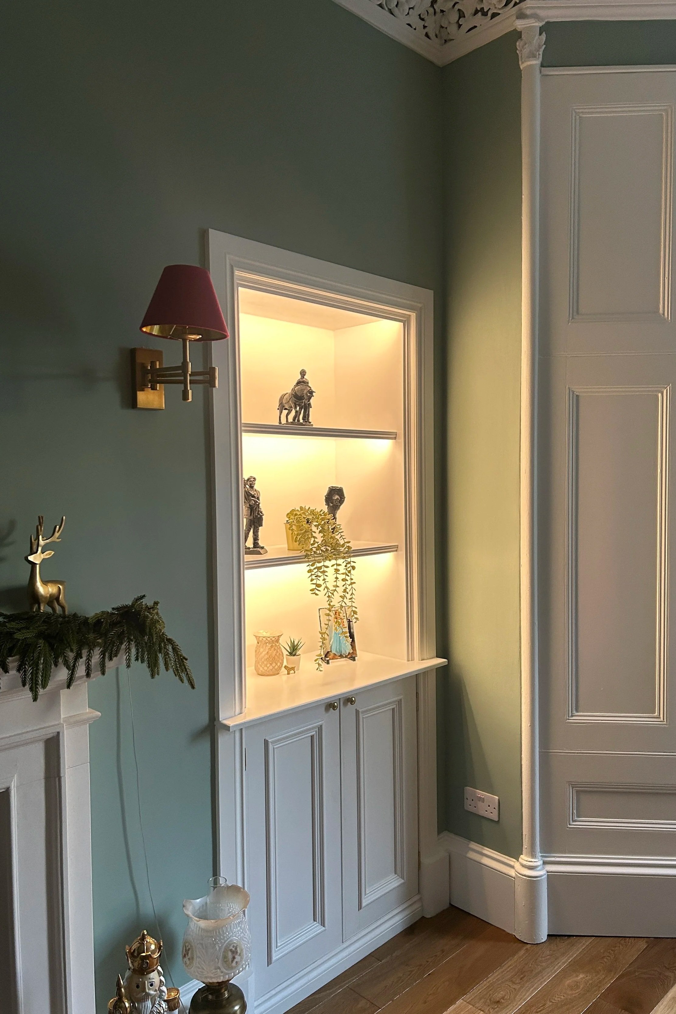 A built-in display cabinet with illuminated shelves holding decorative figurines and plants, positioned against a pastel green wall. To the left, part of a fireplace mantel with festive decorations, and above that, a wall-mounted brass and burgundy lampshade.