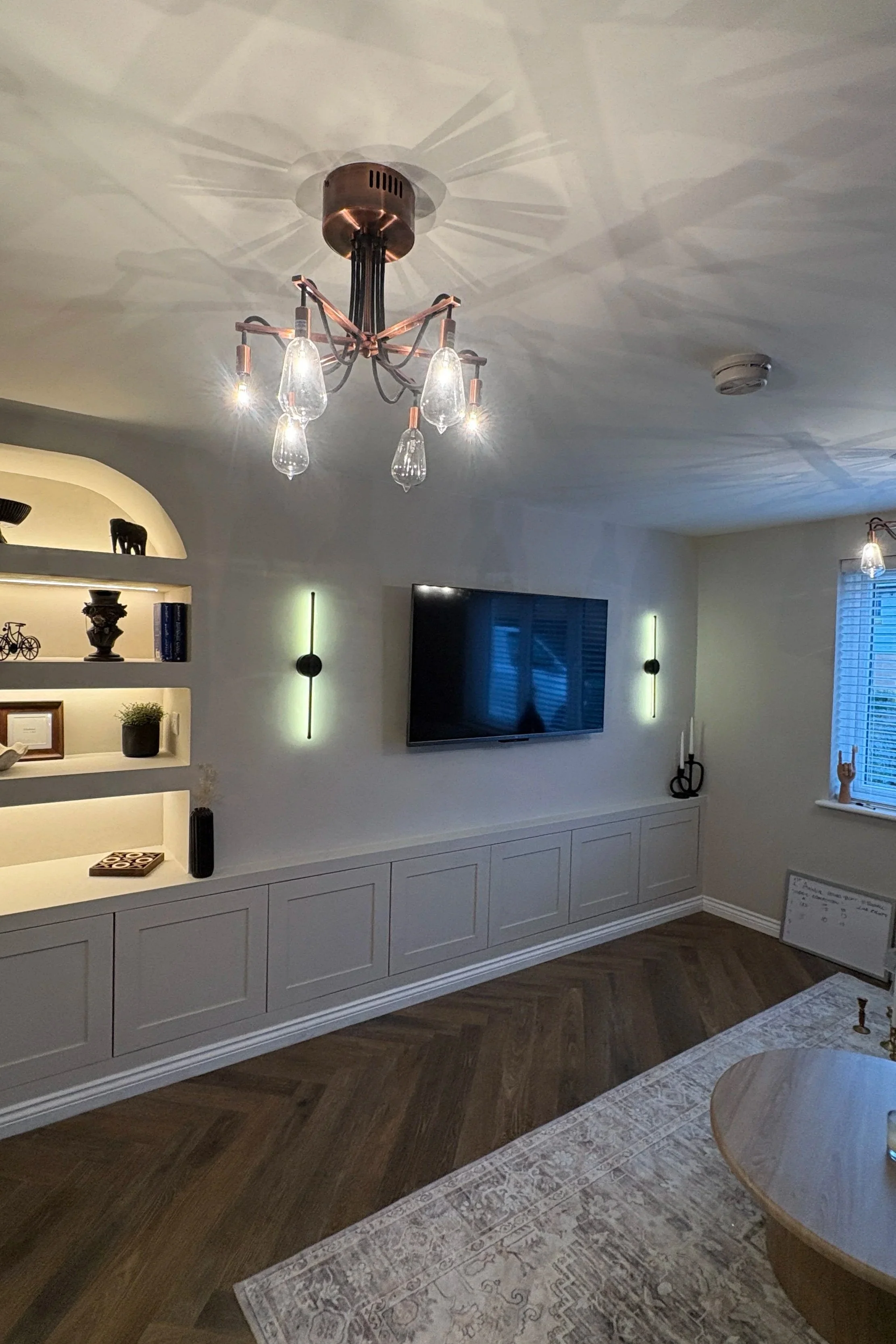 Living room with a wall-mounted flat-screen TV, modern lighting, a built-in white cabinet, and a patterned area rug on wood flooring.