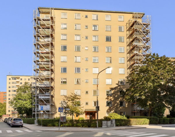 Väggupp byggnadsfas, flerfamiljshus med scaffolding, träd och bilar framför, gata med övergångsställe, blå himmel.