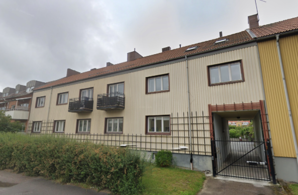 Fasad av flerfamiljshus med fönster, balkonger och en ingång under ett överhäng, omgiven av buskar och en mur.