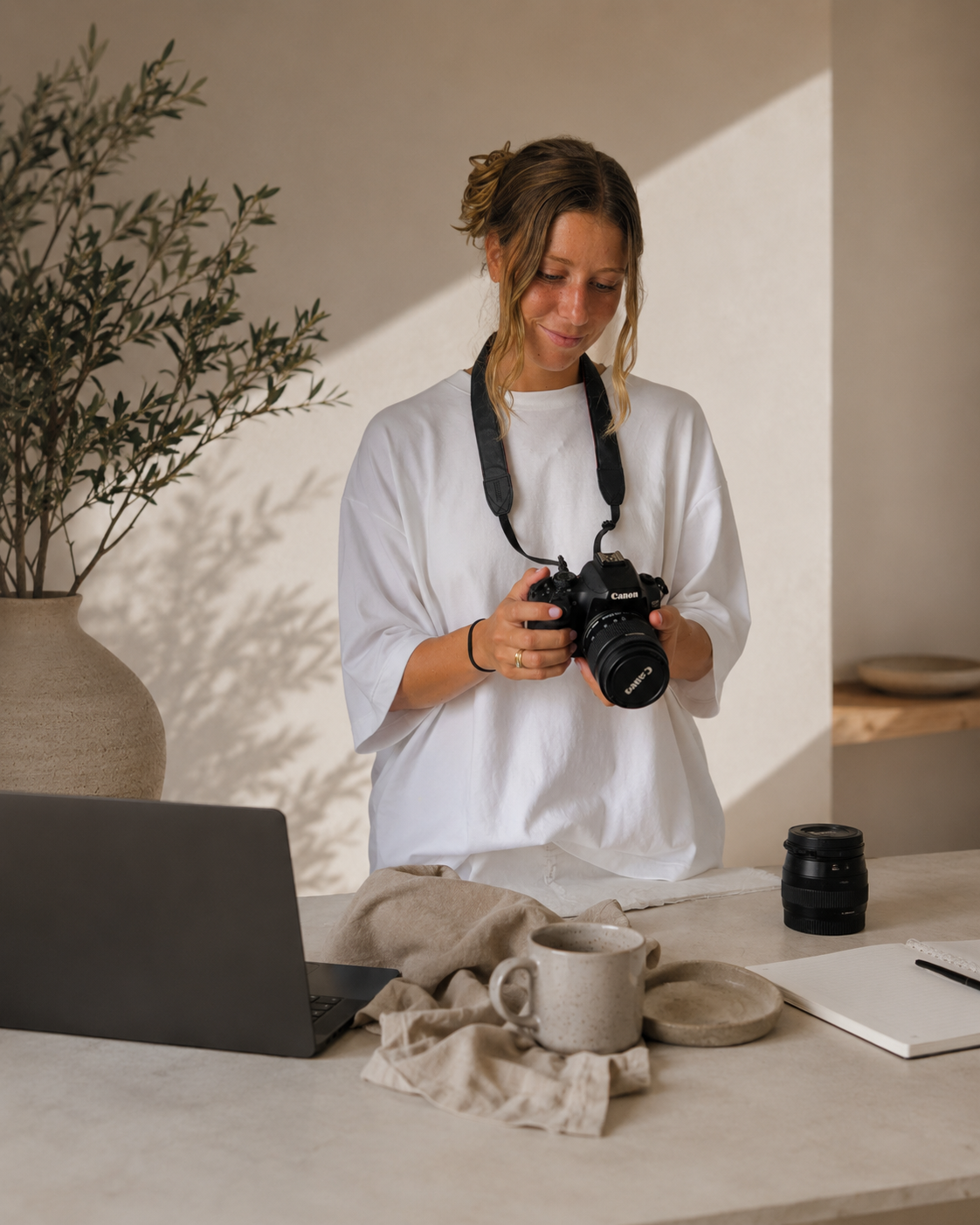 Mujer con cámara fotográfica en una mesa, rodeada de utensilios de cerámica y una laptop en un ambiente iluminado