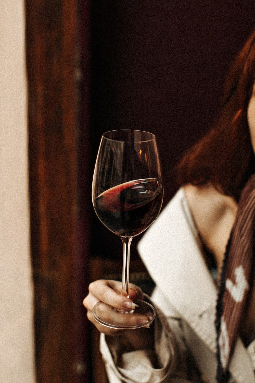 A person holding a glass of red wine indoors, with part of their face and clothing visible.