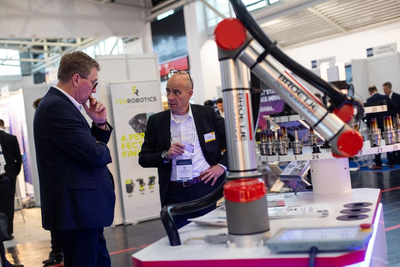 Two men at a robotics exhibition, inspecting a large robotic arm with a busy background of exhibition booths and attendees.