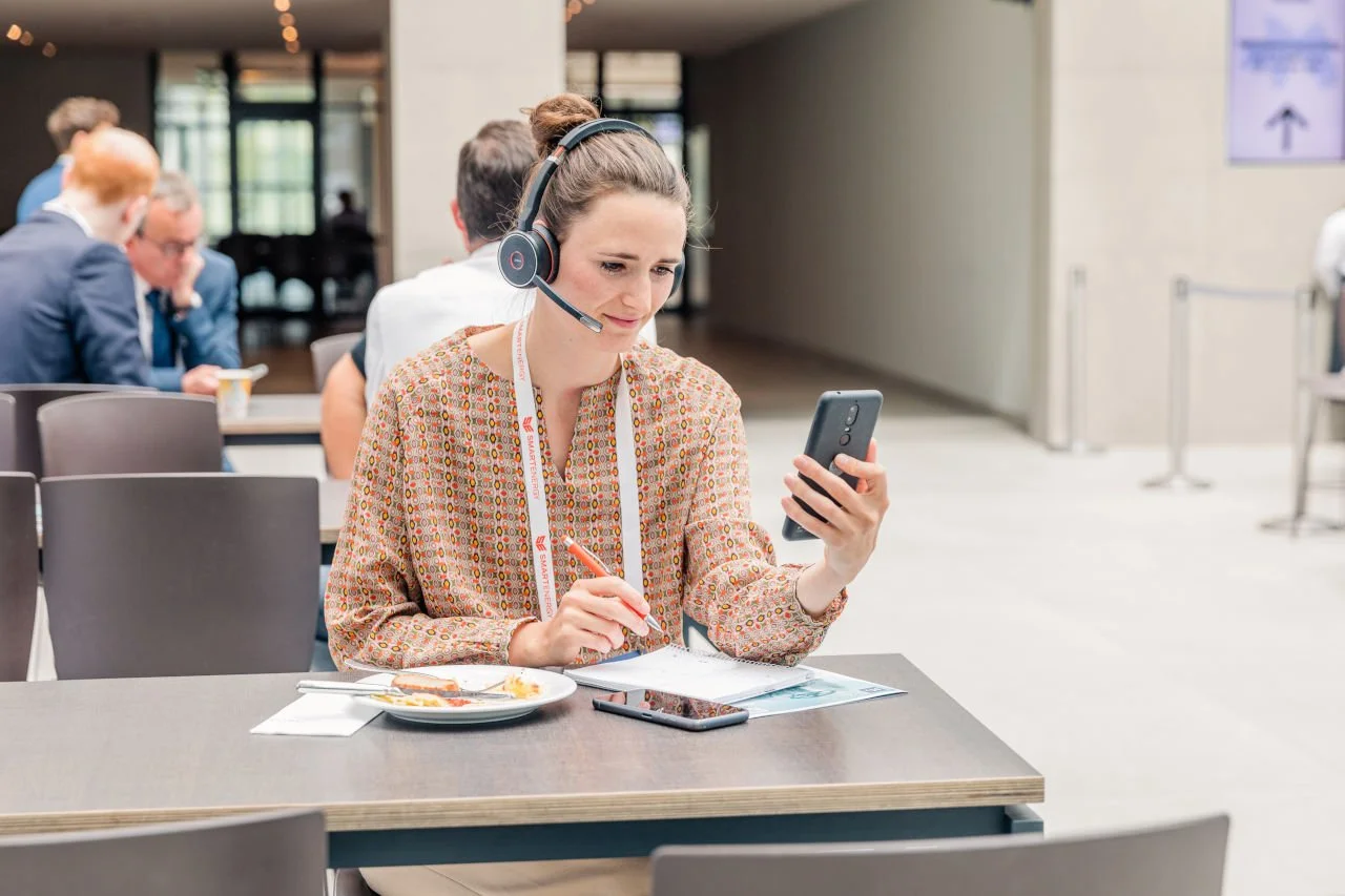 Frau mit Headset und Notizblock arbeitet an einem Tisch in einem Café, während sie auf ihr Smartphone schaut.