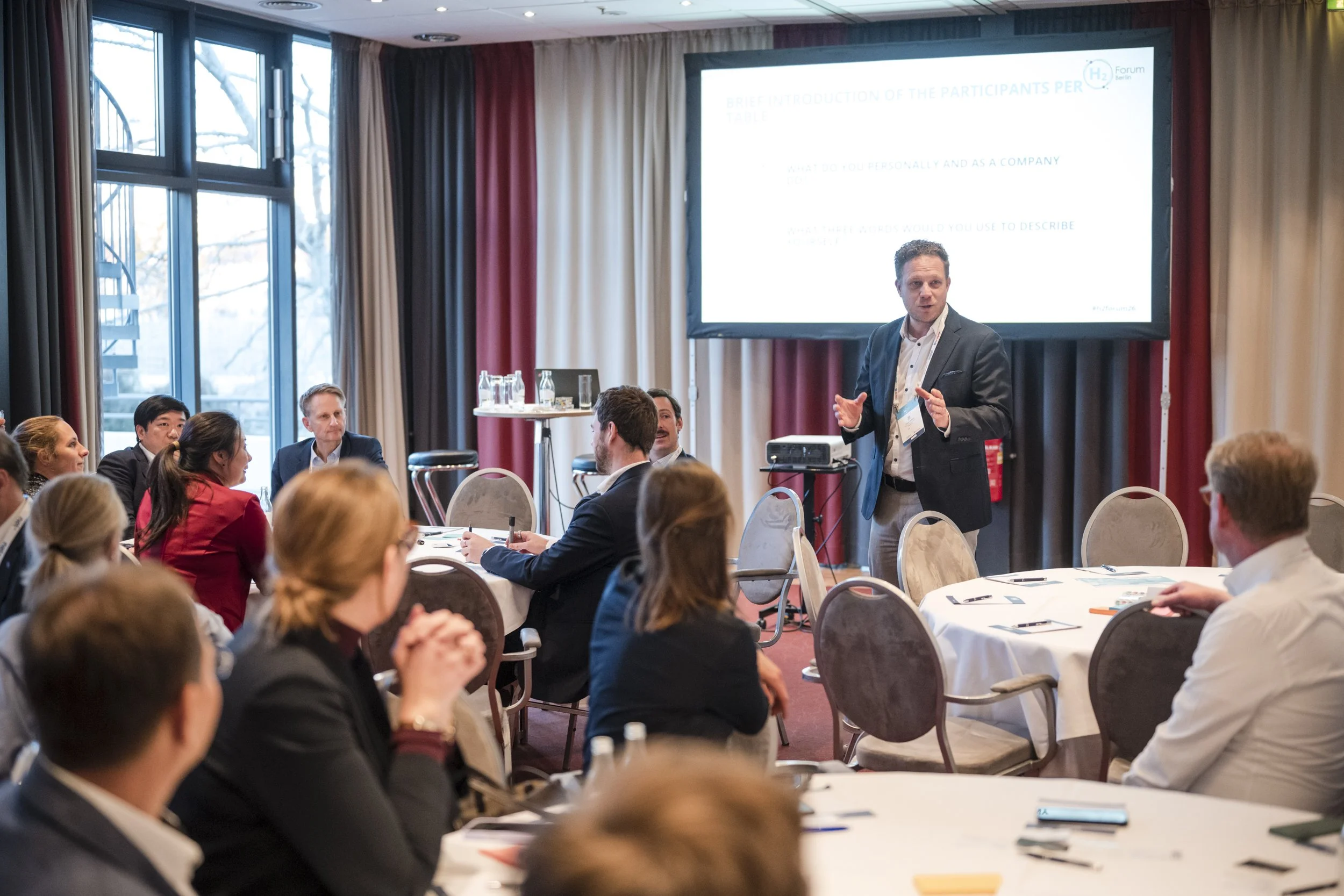 Geschäftsleute bei einem Seminar oder Workshop in einem Tagungsraum, ein Mann steht vorne und präsentiert eine PowerPoint-Folie, der Raum hat große Fenster mit Blick nach draußen, runde Tische mit Schreibmaterialien, Wasser und Gläsern.