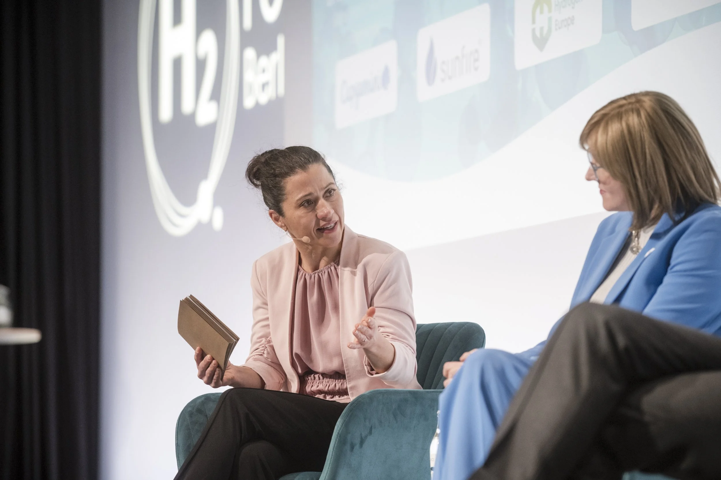 Zwei Frauen im Gespräch bei einer Konferenz, eine spricht, die andere hört zu, im Hintergrund ein Bildschirm mit Logo und Text zur Wasserstofftechnologie.