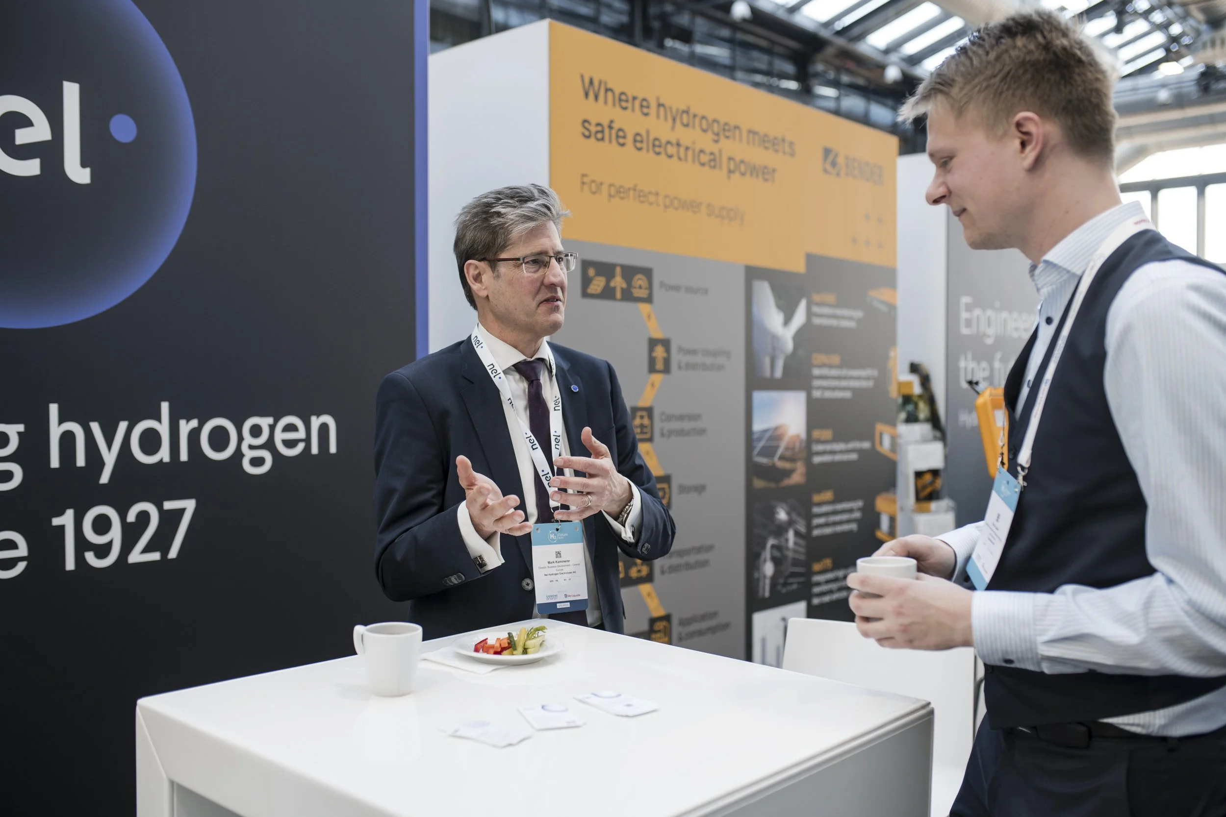 Zwei Männer im Gespräch auf einer Messe, einer erklärt mit Handgesten, während der andere zuhört, im Hintergrund ein Display mit Informationen über Wasserstofftechnologie.