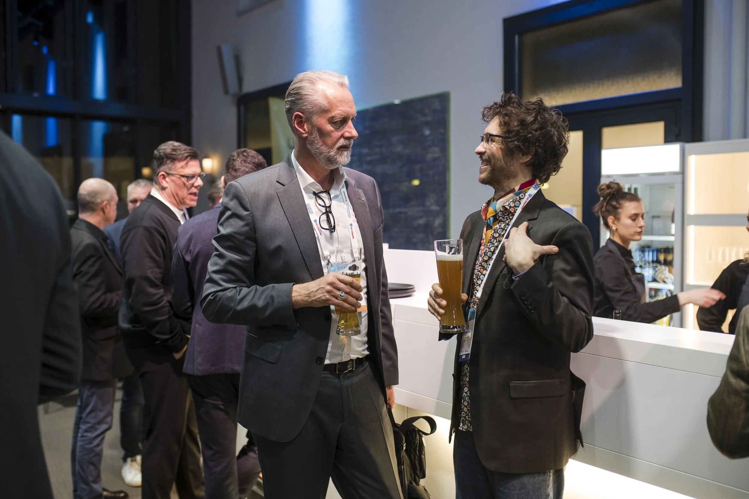 Zwei Männer im Gespräch bei einer Veranstaltung, beide halten Bier. Im Hintergrund sind weitere Personen und eine Bar sichtbar.