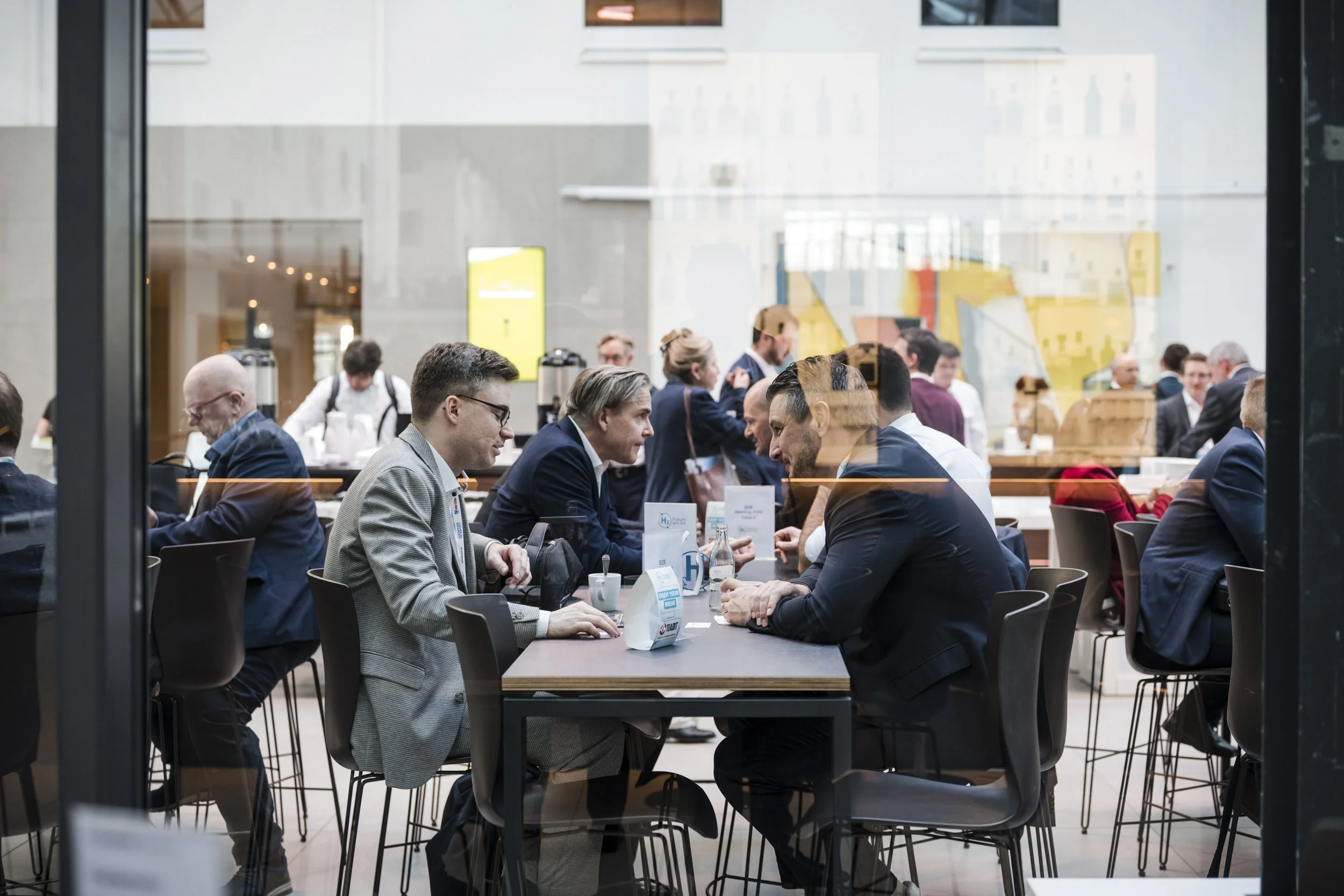 Menschen sitzen an Tischen in einem modernen Café oder Restaurant und führen Gespräche, mit weiteren Personen im Hintergrund, durch eine Fensterfront betrachtet.