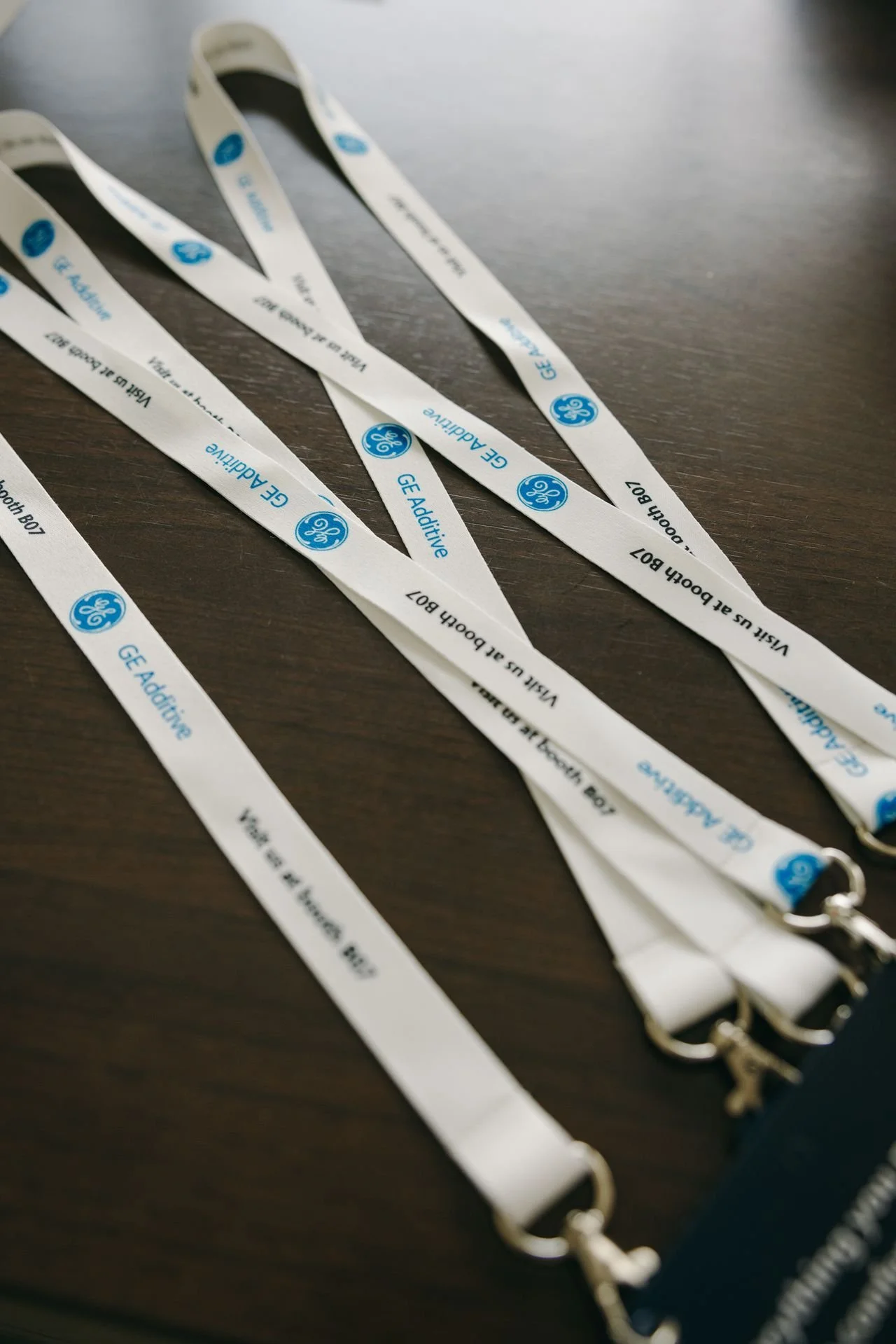Mehrere weiße Lanyards mit blauer Beschriftung und GE Additive Logo, die auf einem dunklen Tisch liegen.
