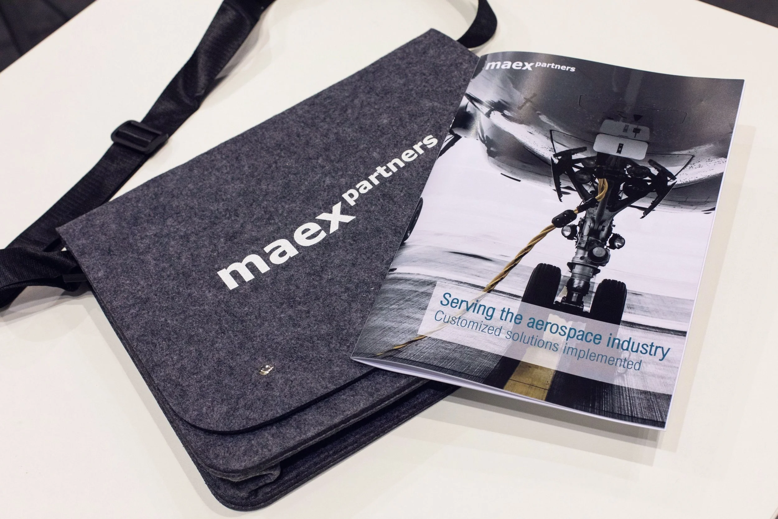 Broschüre mit dem Titel „Serving the aerospace industry“ und einem Bild eines Flugzeuglandebody auf einem Flugzeugträger, auf einem weißen Tisch. Daneben liegt ein grauer Tragetasche mit dem Maex Partners Logo.