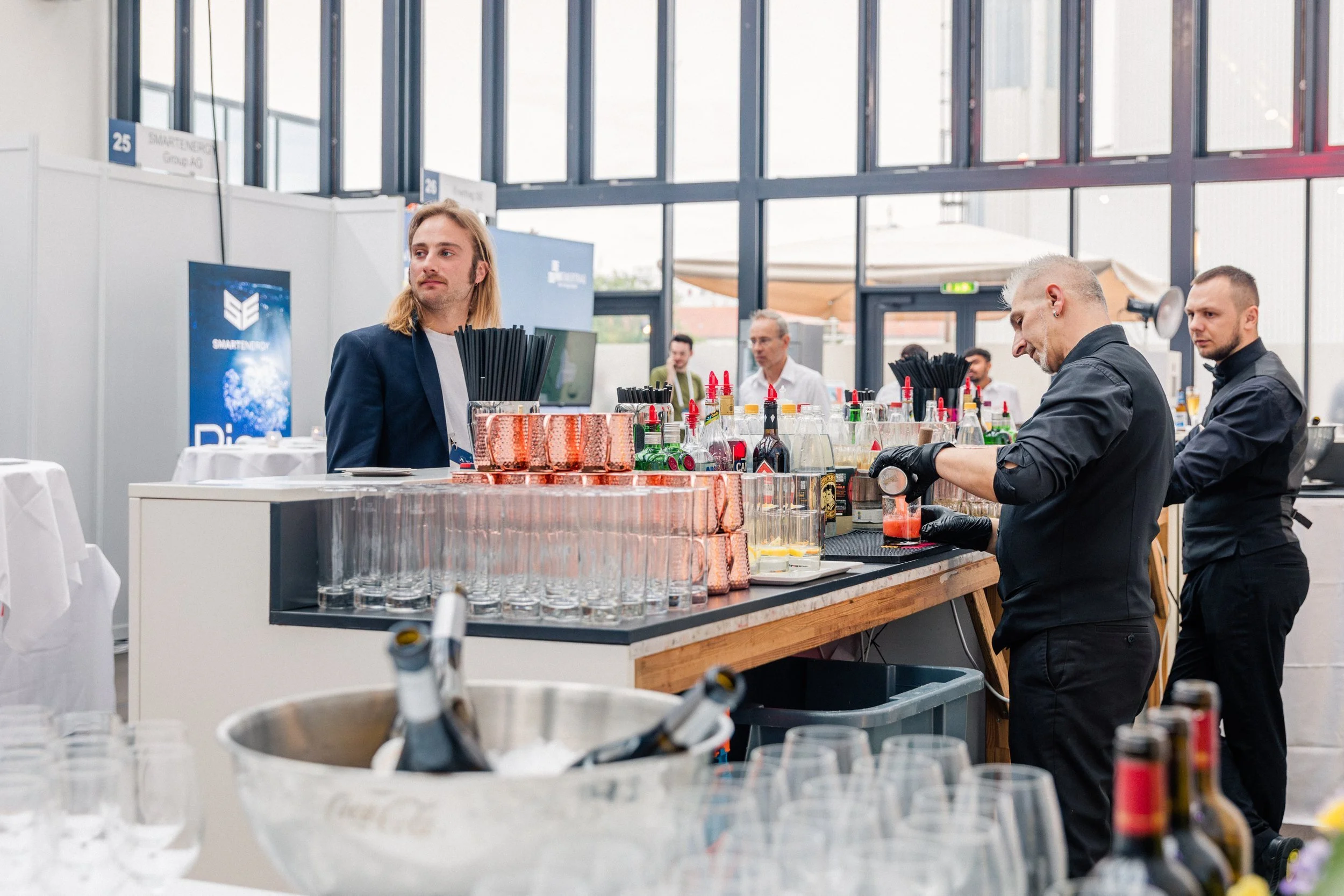 Barkeeper bereitet Getränke an einer Bar bei einer Veranstaltung vor, im Hintergrund stehen Gäste in einem großen, gut beleuchteten Raum.