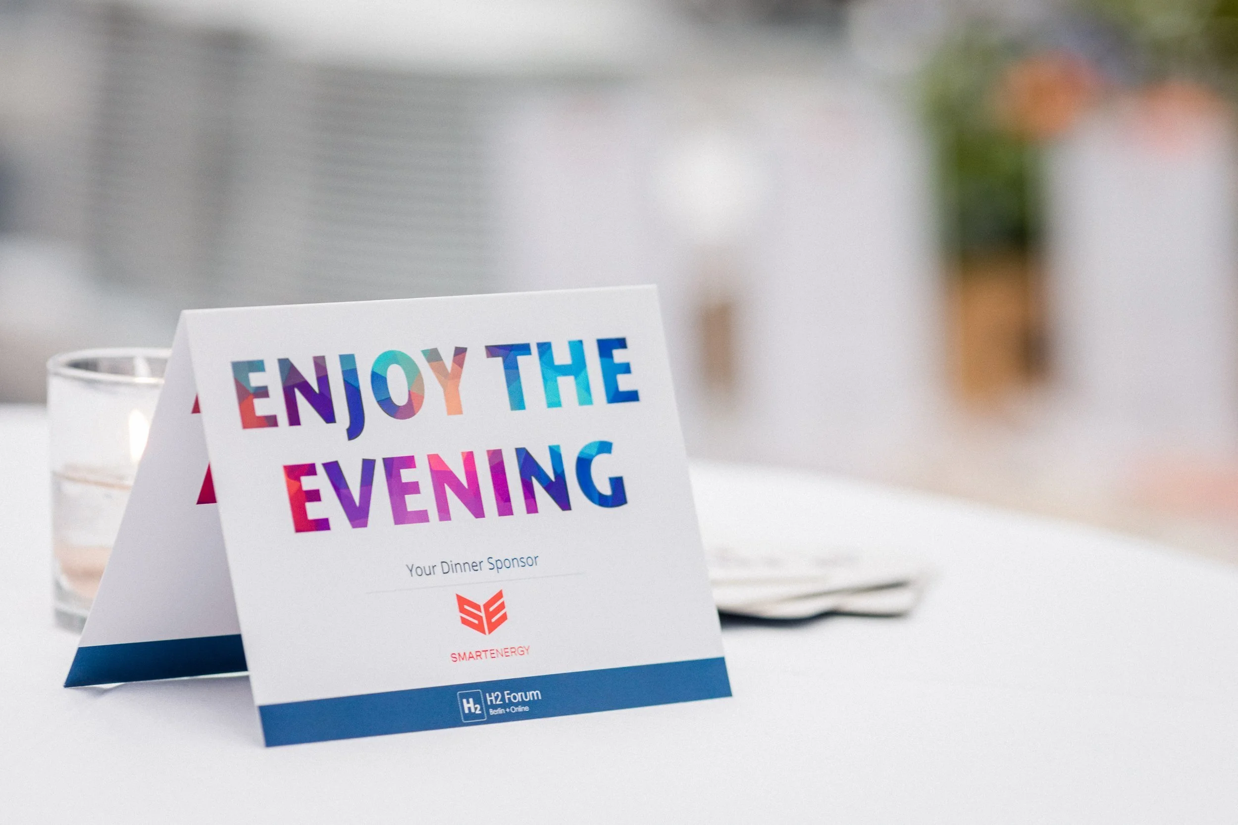 Menükarte mit buntem Schriftzug 'Enjoy the Evening' auf einem weißen Tisch bei einer Veranstaltung, im Hintergrund verschwommenes Dekor.