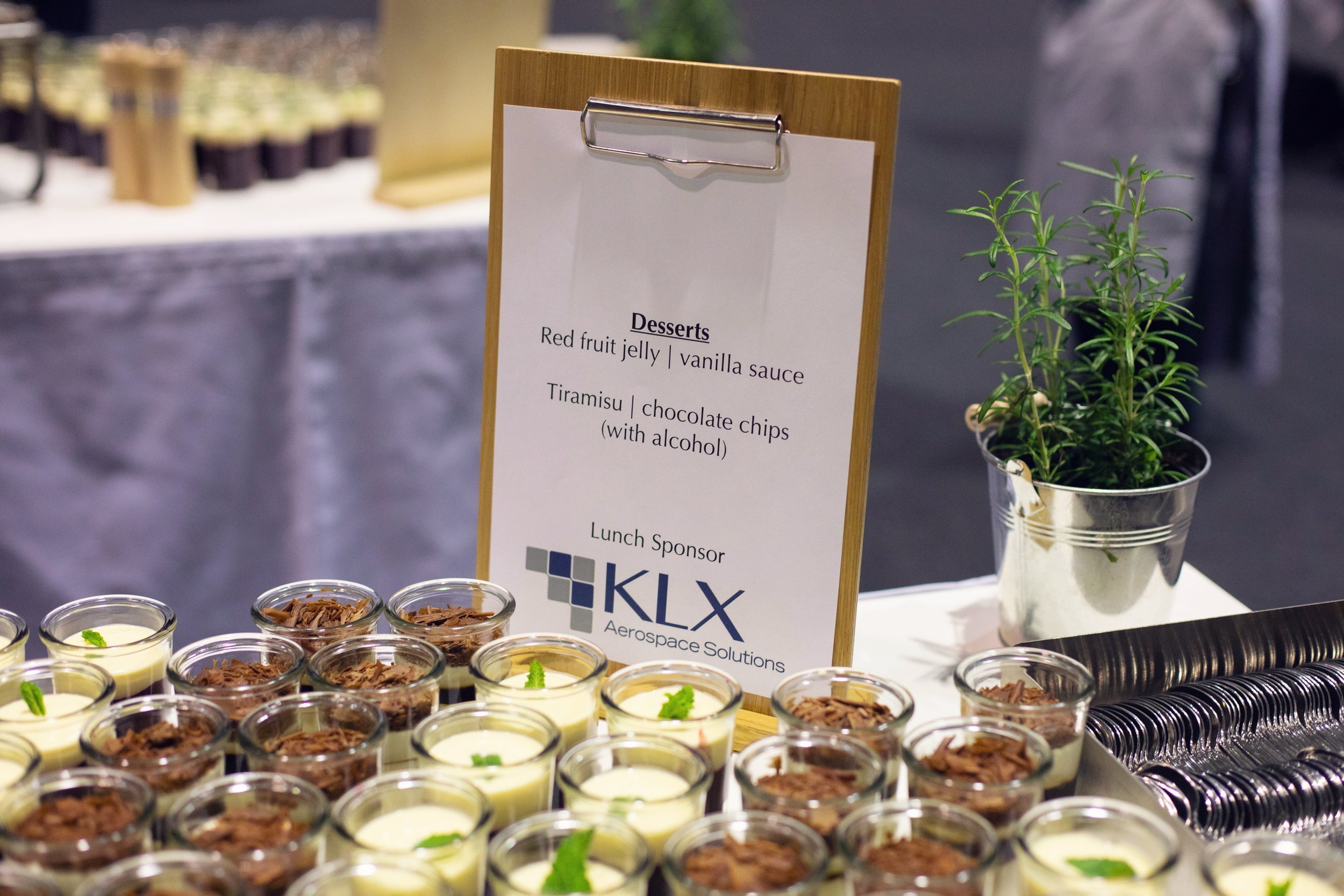 Teller mit kleinen Dessertgläsern, die Schokoladenraspeln und Minzblätter enthalten, auf einem weißen Tisch, neben einem Pappschild mit Speisekarte, der einen Sponsor-Block mit KLX Aerospace Solutions und einer Topfpflanze. Im Hintergrund eine Theke mit weiteren Desserts.