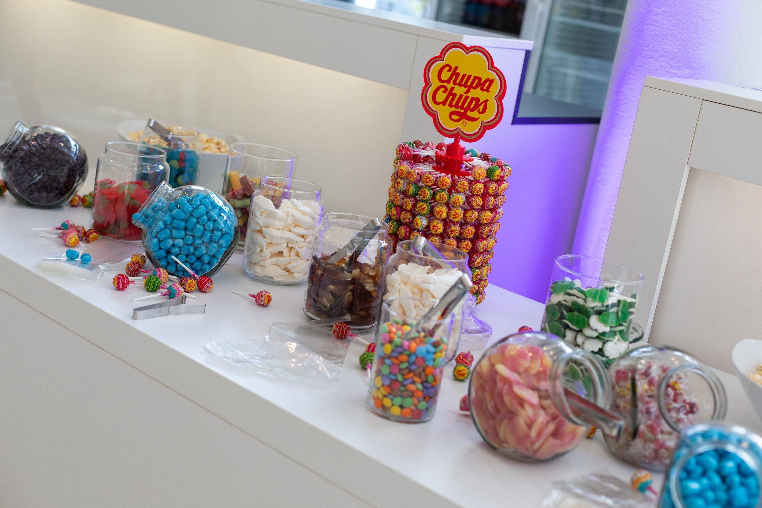 Bunte Süßigkeiten in Gläsern und Tüten, darunter Gummibärchen, Kaubonbons und bunte Schokolinsen, auf einem weißen Tisch in einer dekorierten Umgebung.