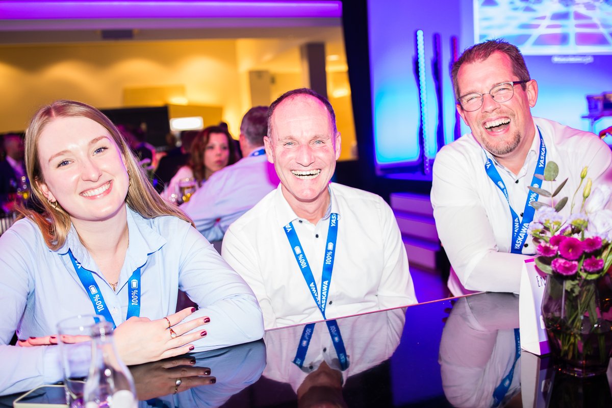 Drei lachende Personen sitzen an einem Tisch auf einer Veranstaltung, mit blauen Lanyards und einem Blumenstrauß auf dem Tisch, im Hintergrund weitere Gäste und eine leuchtende LED-Wand.
