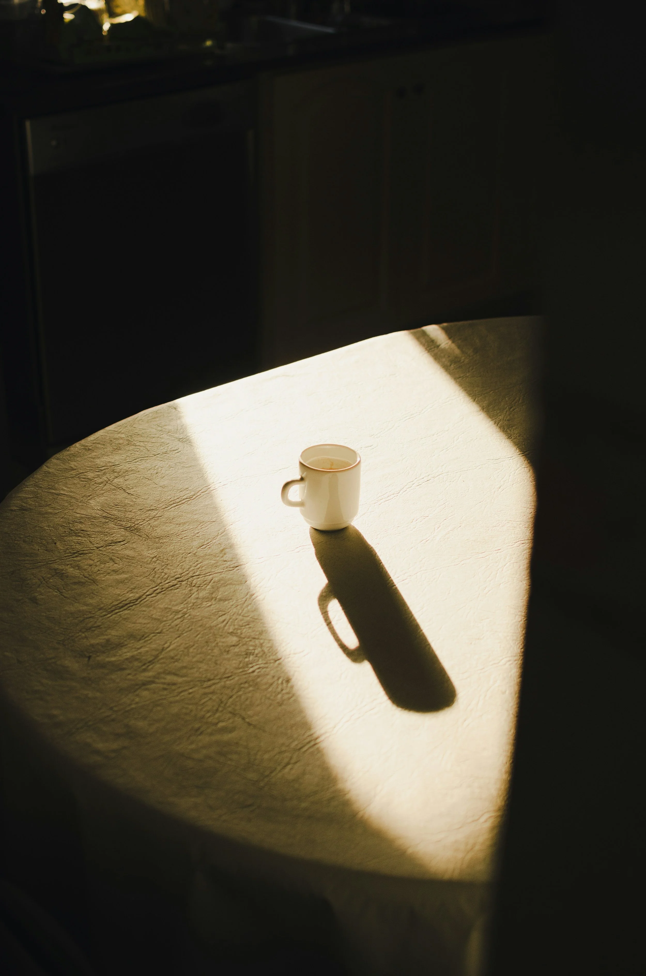 Une tasse blanche posée sur une table, avec une forte lumière naturelle créant une ombre prolongée. En arrière-plan se trouve un meuble sombre.