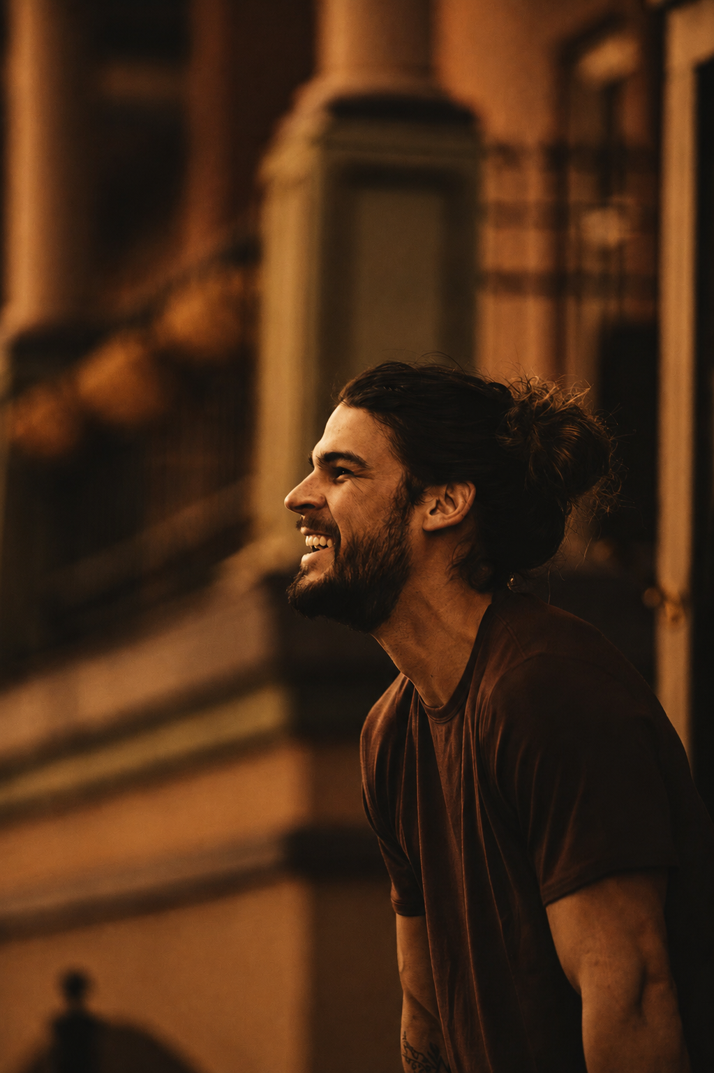 Un homme souriant avec barbe et cheveux longs attachés, portant un t-shirt marron, dans un environnement intérieur chaud et sombre.