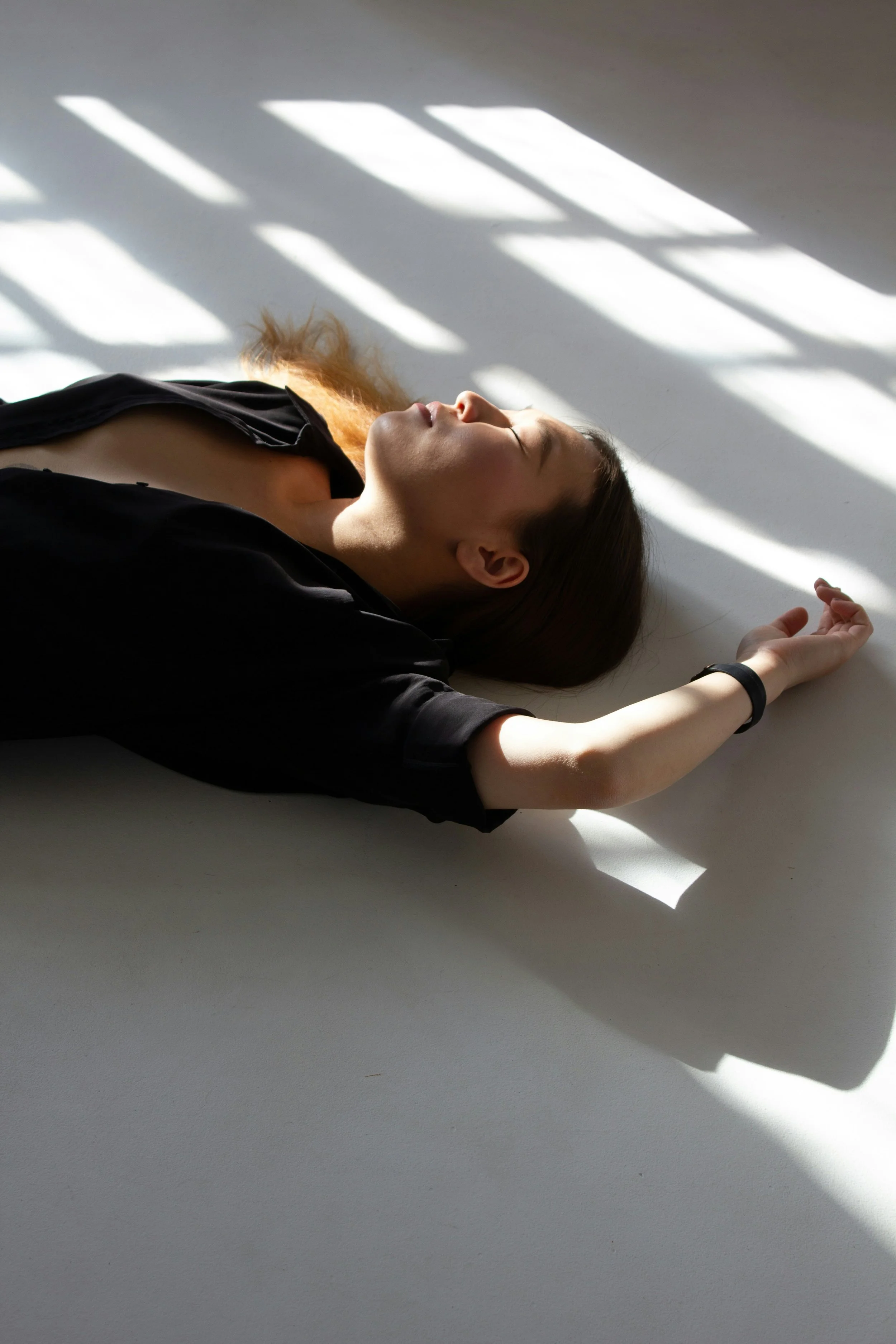 Une femme allongée sur le sol, yeux fermés, avec des rayons de lumière passant à travers des fenêtres, créant des motifs d'ombres sur le mur blanc.