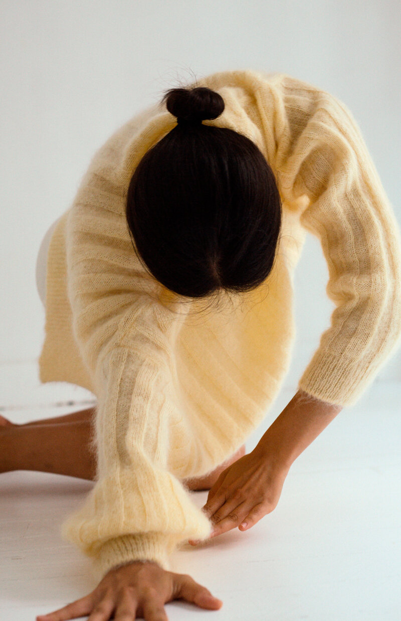 Une femme en pull beige fait une étirement au sol, vue de dessus, ses cheveux sont attachés en bun.