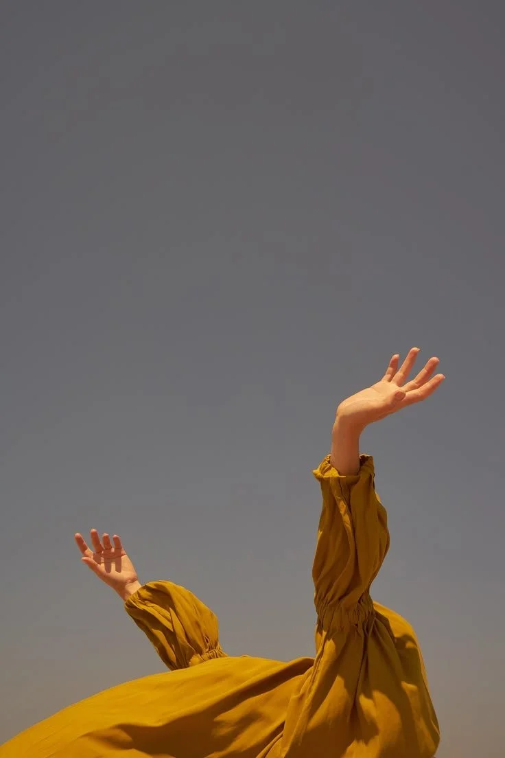 Une personne portant une robe jaune déployée, levant les bras vers le ciel contre un ciel gris.