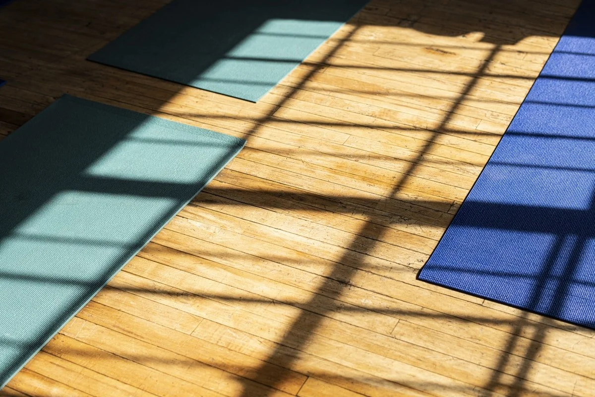 Tapis de yoga en toile turquoise et bleu posés sur un parquet en bois, avec des ombres d'une fenêtre projetant des lignes de lumière.