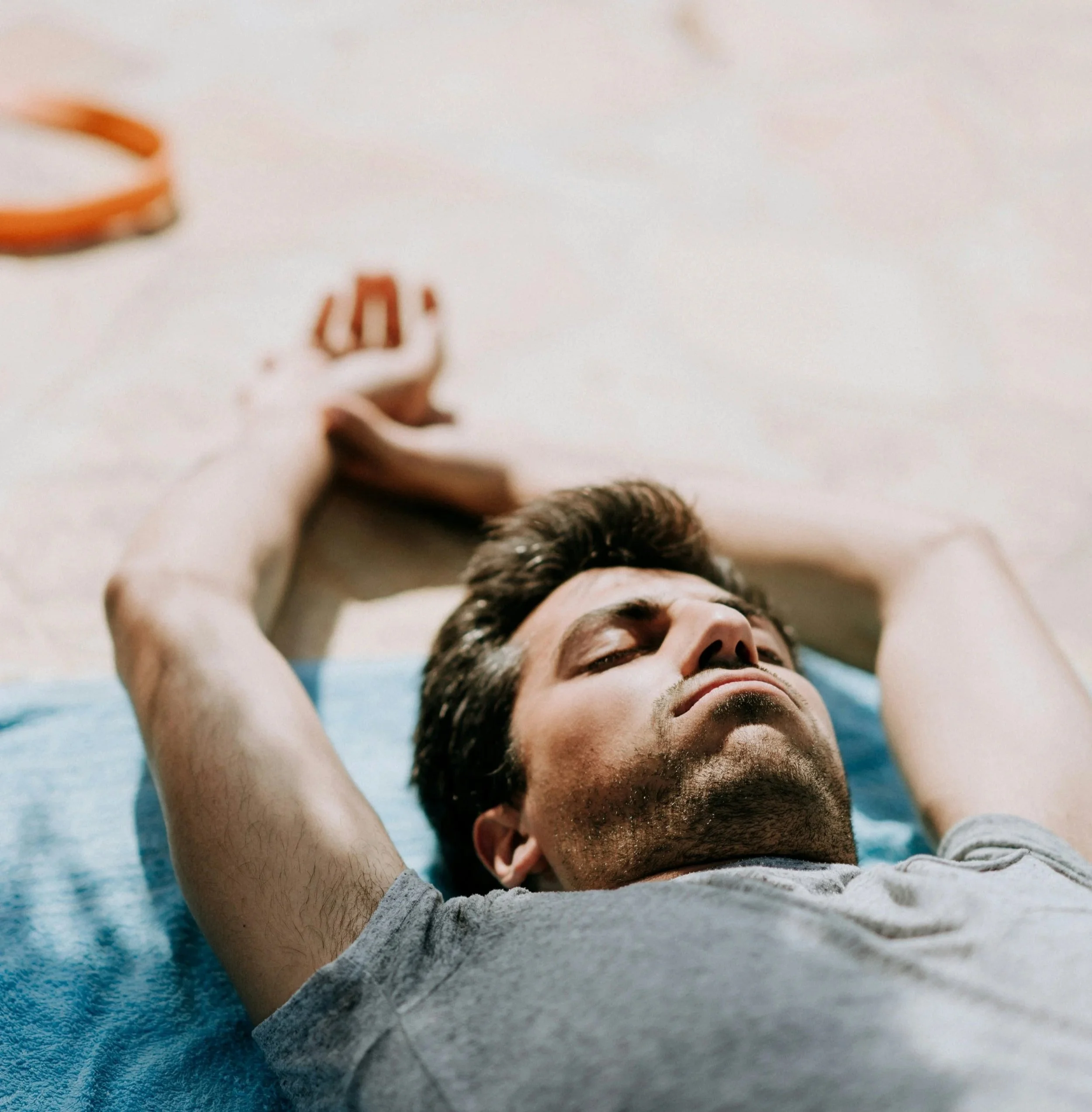 Un jeune homme allongé sur un tapis de sol bleu, les yeux fermés, portant un t-shirt gris clair. Il semble détendu, avec une main derrière la tête. La photo est prise de haut, mettant en valeur son visage reposant et la scène apaisante.